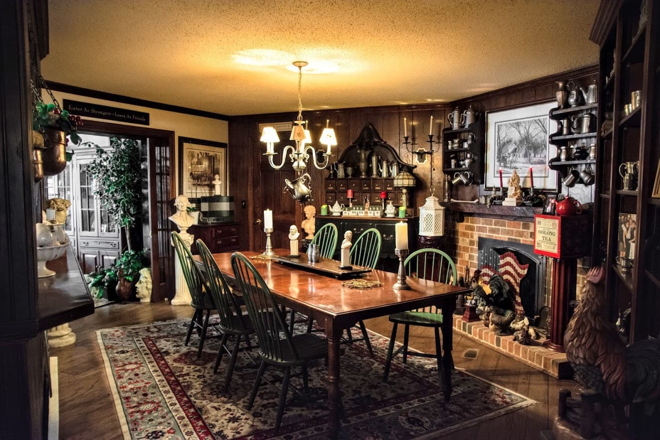 A cozy dining room featuring a wooden table, green chairs, decorative shelves, and a fireplace with various ornaments.