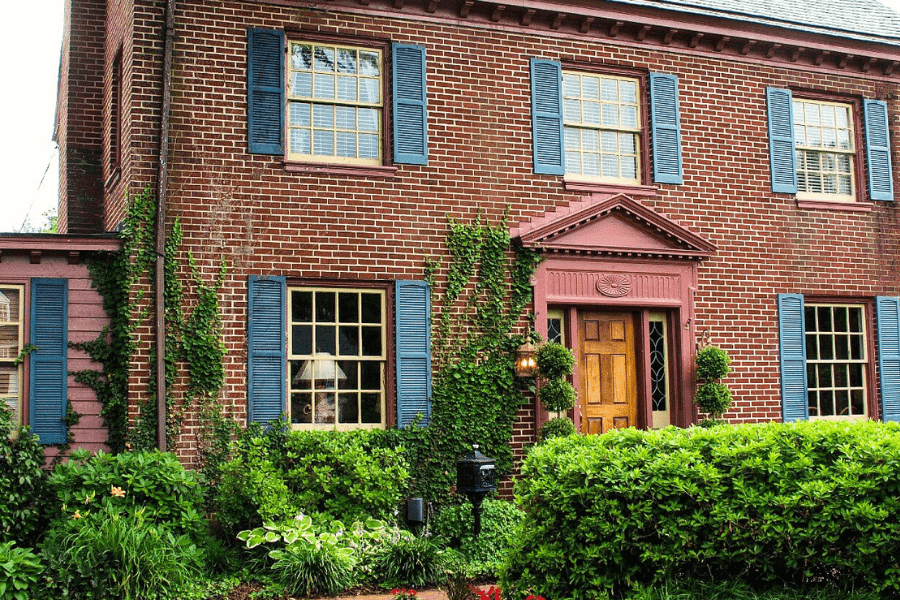 A brick house with blue shutters, a pink entryway, and lush greenery.