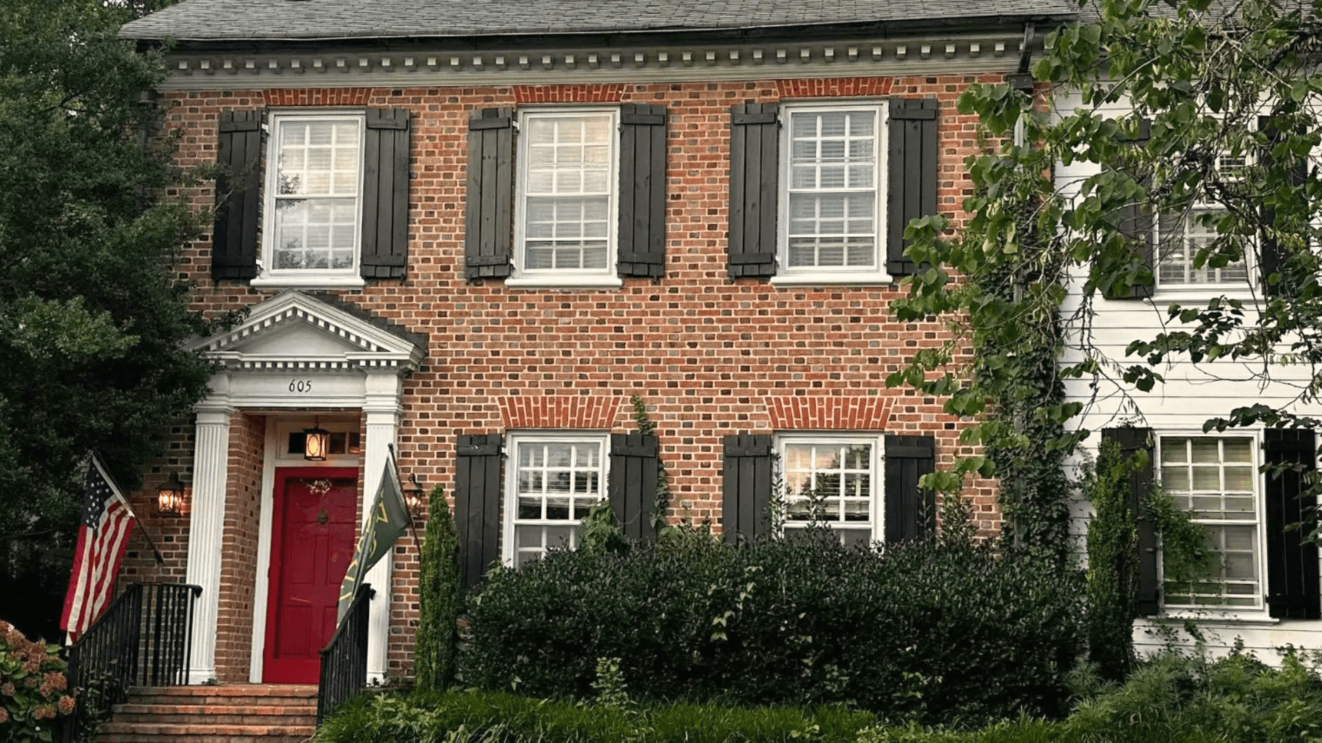 Brick house with a red door and black shutters, surrounded by greenery.
