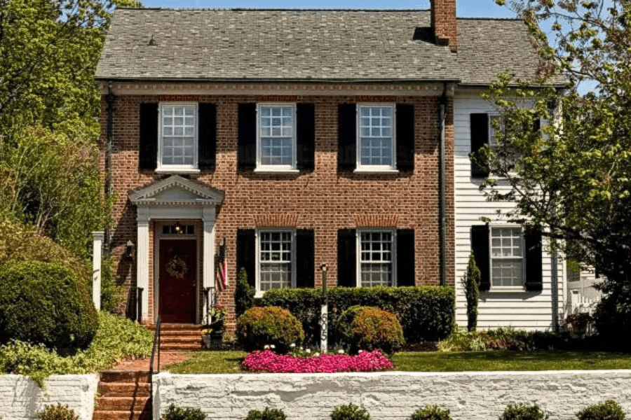 A classic brick and white house with black shutters, surrounded by landscaped greenery and colorful flowers.